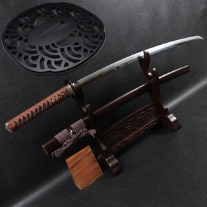 Könnte beinhalten: Ein japanisches Katana-Schwert mit braunem Griff und silberner Klinge, ausgestellt auf einem Holzständer. Der Ständer ist dunkelbraun und hat dekorative Schnitzereien. Ein schwarzer, kreisförmiger Handschutz befindet sich im Hintergrund.