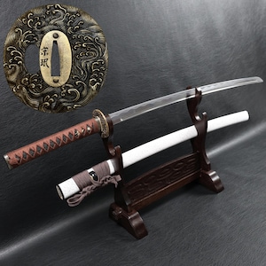 Op de afbeelding: Een Japanse katana-zwaard met een bruin handvat en een witte schede, tentoongesteld op een houten standaard. Het lemmet is zilverkleurig en de pareerstang is goudkleurig. Een decoratief, cirkelvormig metalen stuk met Japanse karakters staat op de achtergrond.
