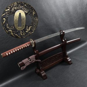 Peut inclure: Épée de style japonais avec un manche et un fourreau marron, présentée sur un support en bois. La lame argentée est complétée par une garde en bronze ornée d'un motif de dragon. Une arme traditionnelle.