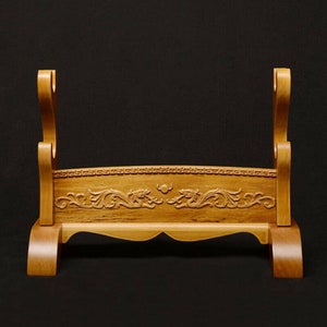 May include: A wooden sword stand with a carved dragon design. The stand is made of light brown wood and has two slots for swords.