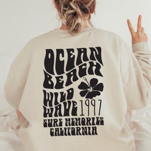May include: A beige sweatshirt with a black graphic print on the back. The graphic says "Ocean Beach Wild Wave 1997 Sure Memories California" in a retro font with a black hibiscus flower.