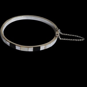 Könnte beinhalten: Ein silbernes Armreif-Armband mit einem geometrischen Muster in Schwarz und Weiß. Das Armband hat eine kleine Kette, die am Verschluss befestigt ist.