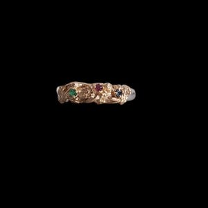 May include: A gold ring with a delicate design featuring four small gemstones: a green emerald, a red ruby, a clear diamond, and a blue sapphire.
