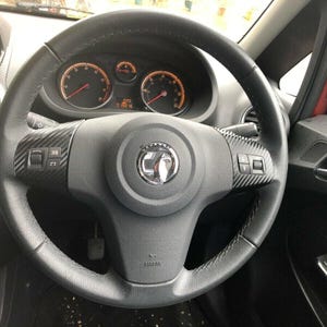 May include: A black leather steering wheel with carbon fibre accents and integrated controls. The dashboard features gauges with orange needles and a digital display. The centre of the wheel displays a silver emblem, and the airbag is labelled.
