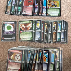 May include: A collection of Dragon Ball Z trading cards arranged in three horizontal rows. Each card features character illustrations, text, and game statistics. The cards are encased in clear plastic protective sleeves, displaying various colors and designs.