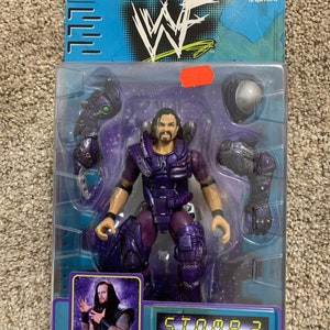 May include: A purple and black action figure of the Undertaker from the WWE S.T.O.M.P. 3 Space Domination line. The figure is wearing a futuristic suit with a helmet and has a removable arm accessory.