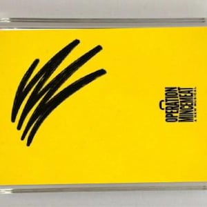 May include: A rectangular clear acrylic magnet featuring a bright yellow background. On the left, there are four thick black hand-drawn diagonal strokes. On the right, the black text reads OPERATION MINCEMEAT A NEW MUSICAL with a small briefcase icon.