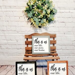 May include: Three wooden signs with the text "this is us our life. our story. our home." in black lettering. The signs are displayed on a wooden stand in front of a white brick wall with a green wreath hanging above.