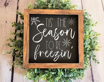 May include: A wooden framed sign with a black background and white text that reads "Tis' the Season to be Freezin'". The sign is decorated with snowflake designs.