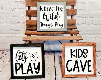 May include: Three wooden signs with white backgrounds and black lettering. The top sign says "Where the Wild Things Play". The middle sign says "lets PLAY" with a small heart. The bottom sign says "KIDS CAVE".