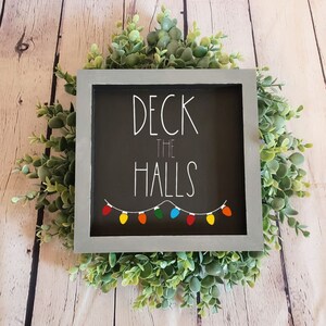 May include: A gray wooden sign with the text "Deck the Halls" in white lettering. The sign has a black background and a string of colorful lights hanging below the text.