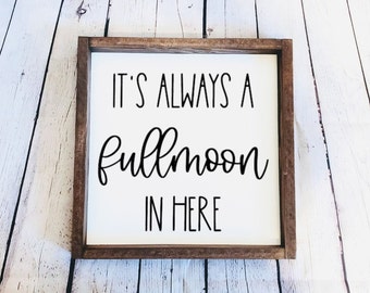 May include: A white wooden sign with a brown frame, featuring the text "It's always a full moon in here".