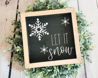 May include: A black framed sign with a white background and the text "Let it snow" in white cursive lettering. There are three white snowflake designs on the sign.