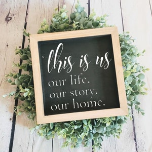 May include: A black framed sign with white text that reads "This is us our life. our story. our home." The sign is surrounded by a wreath of green leaves.