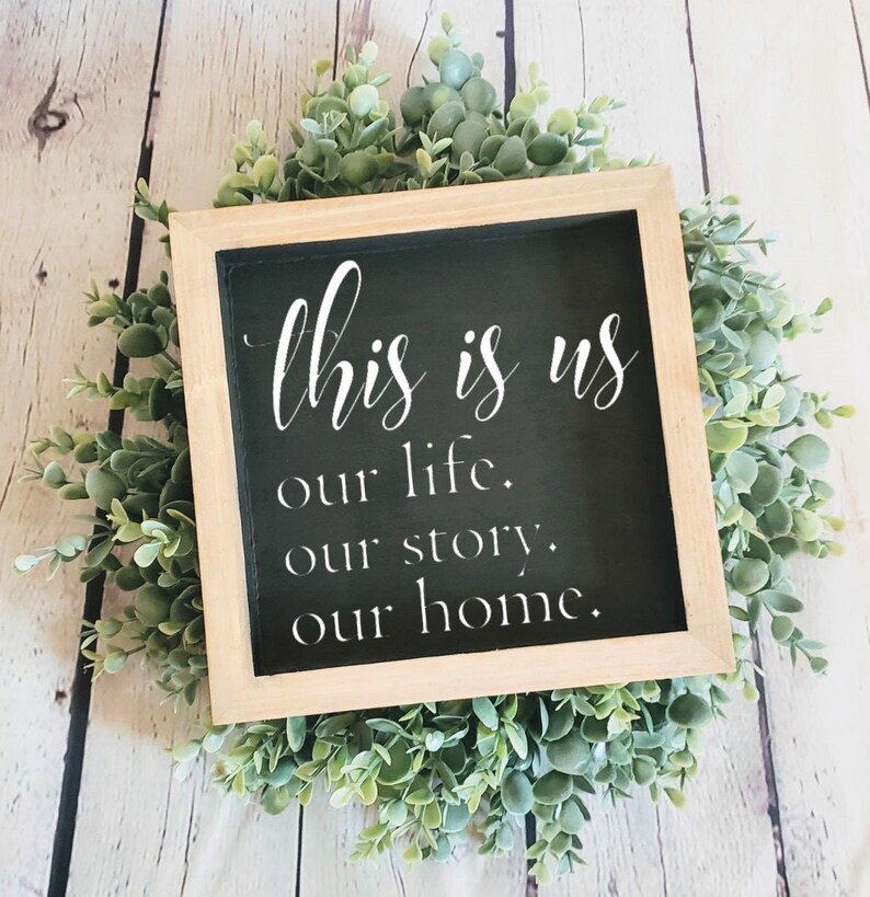 May include: A black framed sign with white text that reads "This is us our life. our story. our home." The sign is surrounded by a wreath of green leaves.