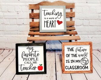 May include: Three wooden signs with black lettering and red accents. The top sign reads "Teaching is a work of HEART" with a red heart. The middle sign reads "my favorite PEOPLE CALL ME Teacher" with a red apple. The bottom sign reads "THE future OF THE world IS IN MY CLASSROOM" with a globe.