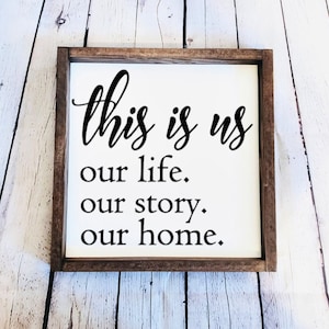May include: A wooden framed sign with the text "this is us our life. our story. our home." in black cursive font on a white background.
