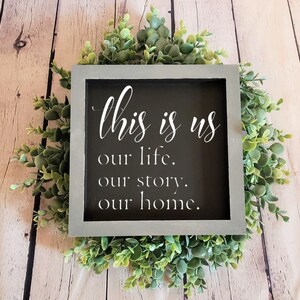 May include: A gray framed sign with the text "This is us our life. our story. our home." in white lettering on a black background. The sign is surrounded by a wreath of green foliage.