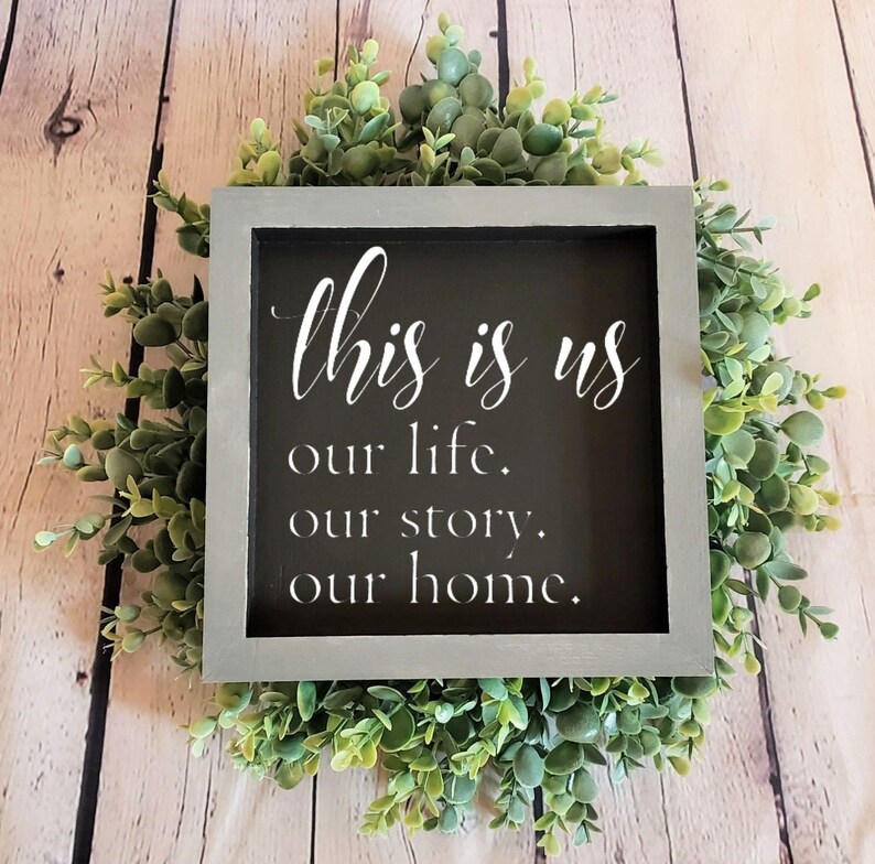May include: A gray framed sign with the text "This is us our life. our story. our home." in white lettering on a black background. The sign is surrounded by a wreath of green foliage.