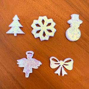 May include: Five iridescent Christmas ornaments: a tree, a snowflake, a snowman, an angel, and a bow. Each ornament has a glittery, textured surface with a mix of pastel colors, including pink, yellow, and blue. The ornaments are arranged on a wooden surface.