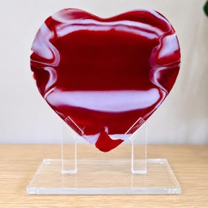 May include: A heart-shaped decorative glass piece with a vibrant red and white swirled design. The glass is displayed on a clear acrylic stand, showcasing its unique shape and color. The piece is a decorative item.