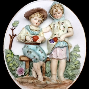 May include: A porcelain wall plaque featuring two children, a boy and a girl, sitting on a bench in a garden setting. The boy is wearing a blue and white outfit and holding an apple. The girl is wearing a pink and white dress and holding a bunch of grapes. The plaque is decorated with flowers and leaves.