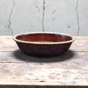 May include: A round ceramic bowl with a deep brown, glossy interior and exterior finish. The rim features a contrasting, textured beige and cream speckled glaze that creates a distinct border around the edge of the circular vessel.
