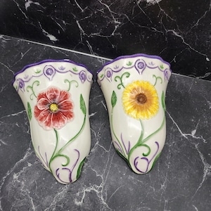 May include: Two white ceramic wall vases with floral designs. One vase features a red flower, the other a yellow sunflower. Both have purple trim and green vines. Decorative home decor.