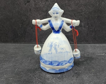 May include: A ceramic figurine of a woman in traditional Dutch attire, with a white hat and dress, accented with blue details. The figurine carries two small buckets suspended by red strings. The base of the dress features a blue and white landscape scene.