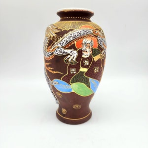 May include: A brown ceramic vase with a dragon and a man in a robe painted on it. The vase has gold accents and is decorated with colorful details.
