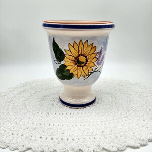 May include: A white ceramic vase with a blue rim and base. The vase is decorated with a hand-painted sunflower and lavender flowers on a light blue background.