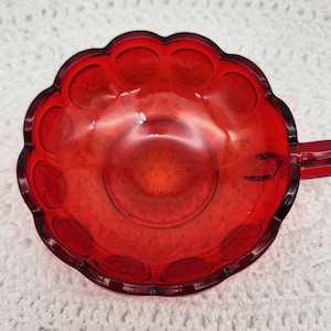 May include: A red glass dish with a scalloped edge and a handle. The dish has a textured surface with small circles.