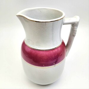 May include: A white and burgundy ceramic pitcher with a gold trim. The pitcher has a handle and a spout for pouring. The burgundy section is a wide band around the middle of the pitcher. The pitcher is likely vintage.
