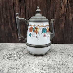 May include: Antique white ceramic teapot with a metal lid and handle. The teapot is decorated with colorful autumn leaves. The handle and lid are a dark gray color. The teapot is sitting on a gray surface.