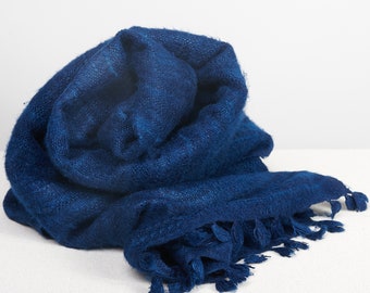May include: A deep blue, soft, fringed scarf made of wool. The scarf is folded and draped on a white surface.