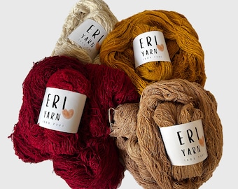 Peut inclure: Quatre écheveaux de fil Eri de différentes couleurs : beige, jaune moutarde, rouge et marron. Chaque écheveau porte une étiquette blanche avec le texte « ERI YARN 100 % PURE » et un petit cœur.
