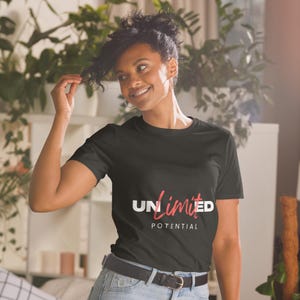 May include: Black t-shirt with the words "UNlimited POTENTIAL" in white and red script. The shirt is short-sleeved and has a crew neck. The model is wearing blue jeans and a black belt.