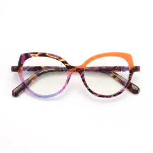 Puede incluir: Gafas de ojo de gato con un patrón de carey multicolor en tonos morado, marrón y negro, contrastado por un borde superior de montura en color naranja brillante. Las lentes transparentes están montadas en una estructura de forma distintiva.