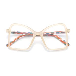 Puede incluir: Gafas de gran tamaño con una montura beige translúcida y una forma de mariposa distintiva. El interior de las patillas presenta un vibrante patrón de cuadros multicolor en tonos rojo, naranja, amarillo, verde, azul y morado.