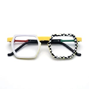 Puede incluir: Gafas con puente y patillas amarillas, con un aro izquierdo blanco sólido y un aro derecho a cuadros blancos y negros. La montura incluye detalles en rojo y verde en las bisagras interiores, creando un aspecto asimétrico y colorido.