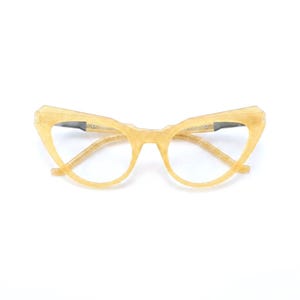 Puede incluir: Gafas de estilo ojo de gato con una montura dorada translúcida y brillante, con un acabado texturizado similar a la purpurina. Las gafas presentan una silueta angular distintiva con esquinas superiores puntiagudas y lentes transparentes.