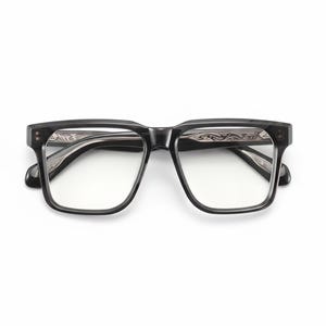 Puede incluir: Gafas rectangulares negras con lentes transparentes. El interior de la montura presenta una incrustación metálica plateada con un intrincado patrón de volutas grabadas visible en el puente y el interior de las varillas.