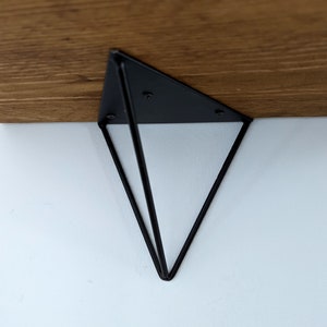 May include: Black metal shelf bracket in the shape of a triangle with a geometric design. The bracket is attached to a wooden shelf.