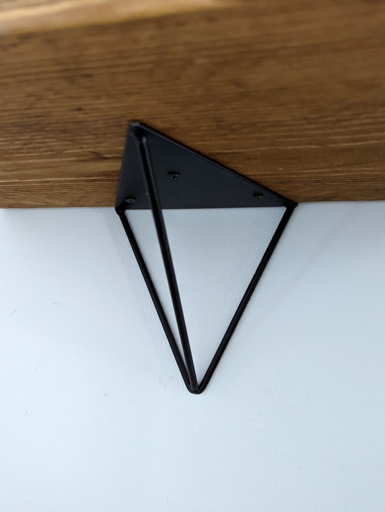 May include: Black metal shelf bracket in the shape of a triangle with a geometric design. The bracket is attached to a wooden shelf.
