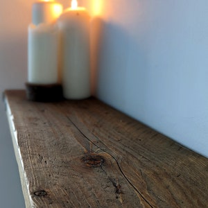 May include: A rustic wooden shelf with a natural brown finish. The shelf is made of reclaimed wood and has a knot in the center. Two white candles are sitting on the shelf.