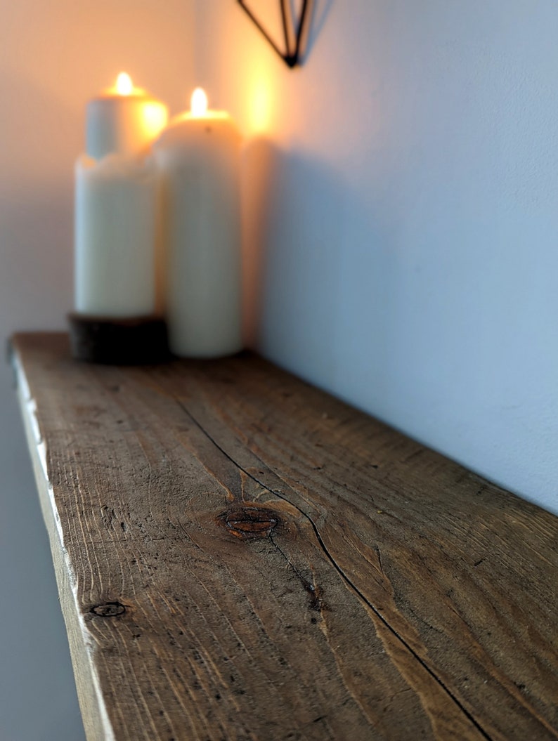 May include: A rustic wooden shelf with a natural brown finish. The shelf is made of reclaimed wood and has a knot in the center. Two white candles are sitting on the shelf.