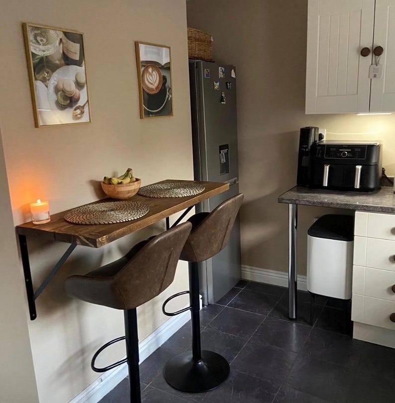 May include: A small kitchen dining area with a wooden table and two brown leather bar stools. The table is supported by black metal brackets. Two framed pictures hang on the wall. A lit candle and a bowl of bananas are on the table.