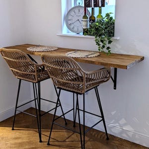 May include: Two woven rattan bar stools with black metal legs sit beneath a wooden countertop. The countertop is attached to a white wall. A window with a clock, wine bottles, and greenery is in the background.