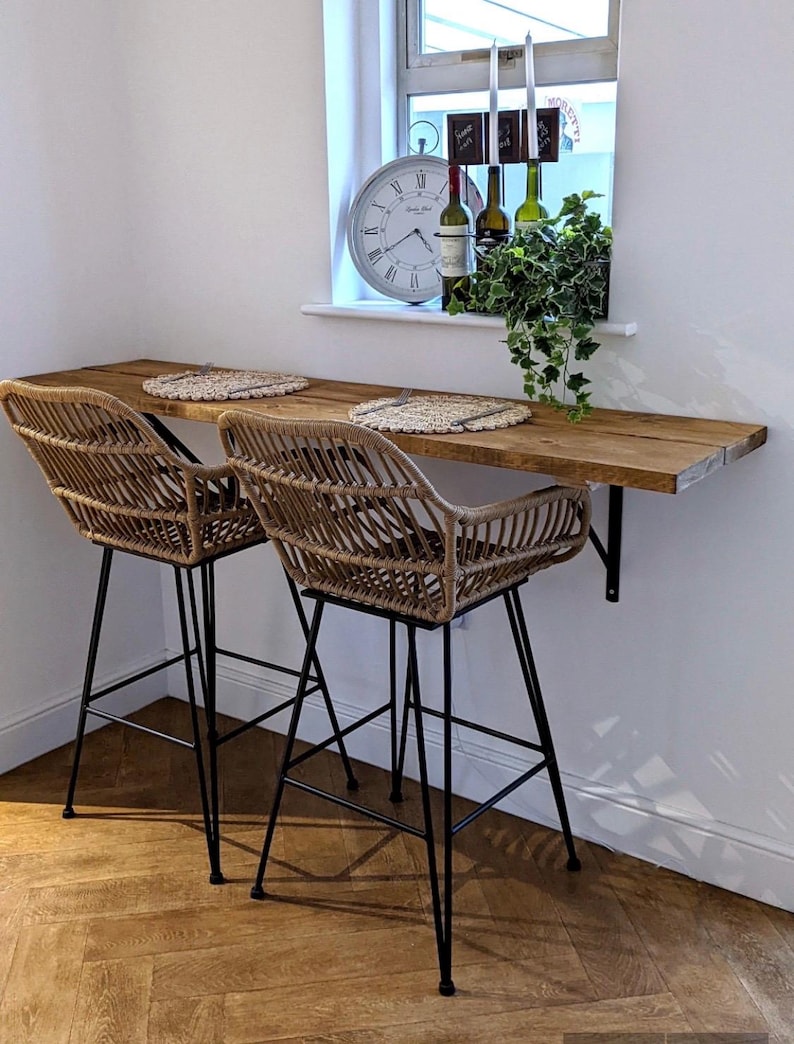 May include: Two woven rattan bar stools with black metal legs sit beneath a wooden countertop. The countertop is attached to a white wall. A window with a clock, wine bottles, and greenery is in the background.