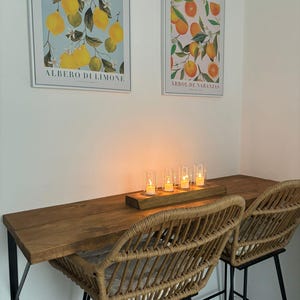 May include: A cozy dining area featuring a wooden table with two woven rattan chairs. Above, two framed botanical prints depict lemons and oranges. A wooden tray holds five lit candles, creating a warm ambiance.